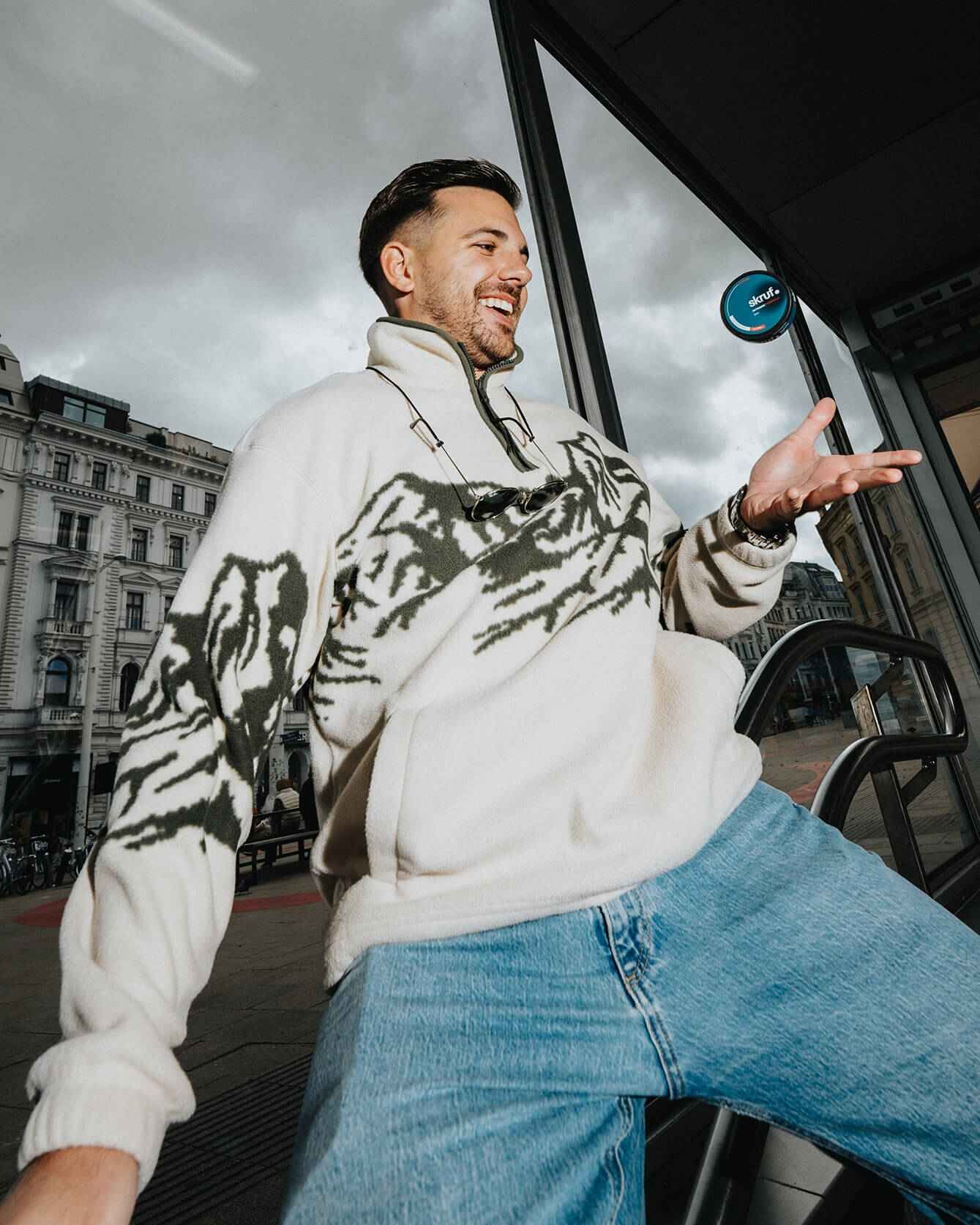 Mann in Fleecejacke mit Skruf-Dose in der Hand vor einer modernen Gebäude-Fassade.