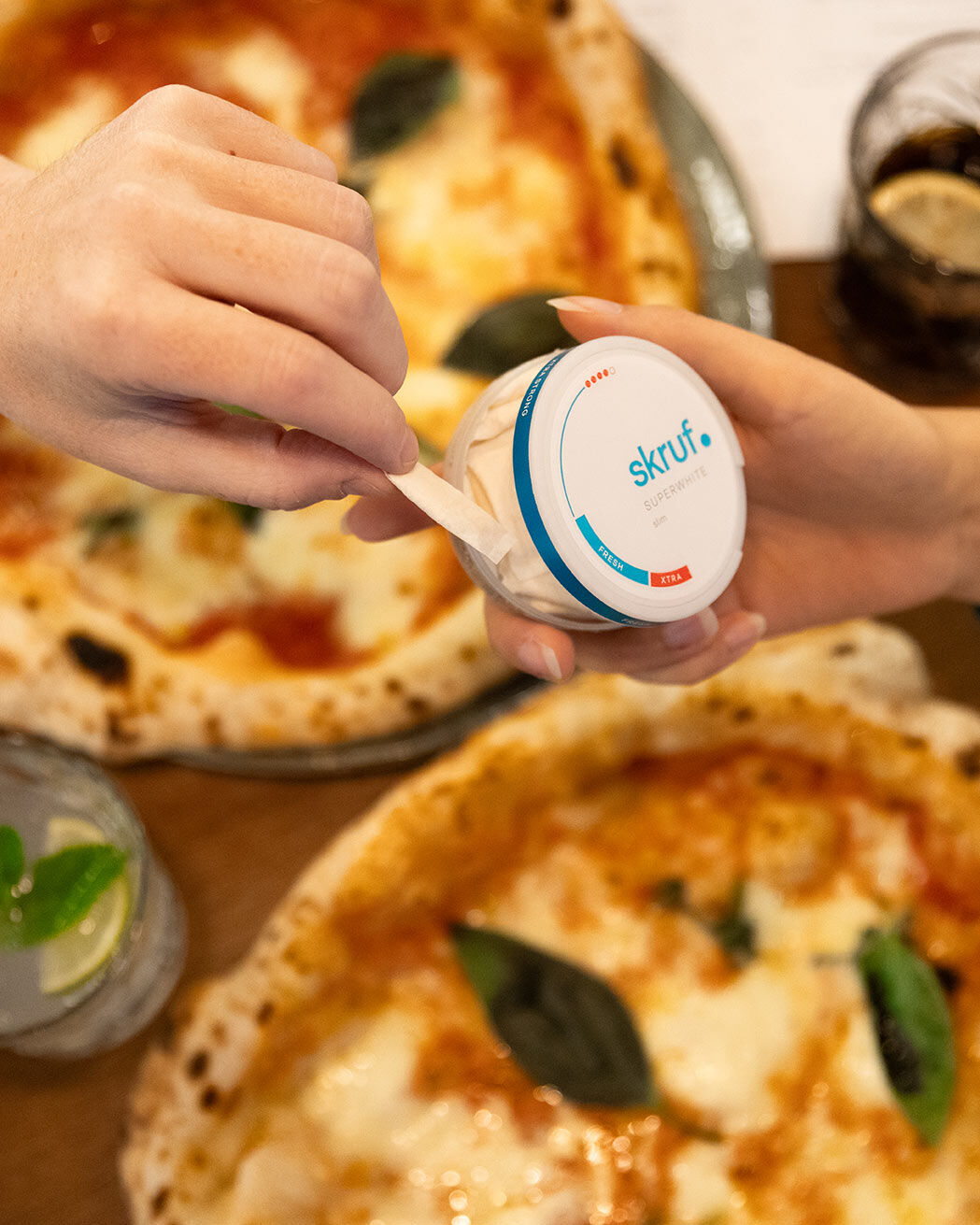 Lifestyle-Shot beim Essen: Eine Person entnimmt einen Beutel aus einer skruf Superwhite Fresh Slim Dose, im Hintergrund stehen zwei frische neapolitanische Pizzen.
