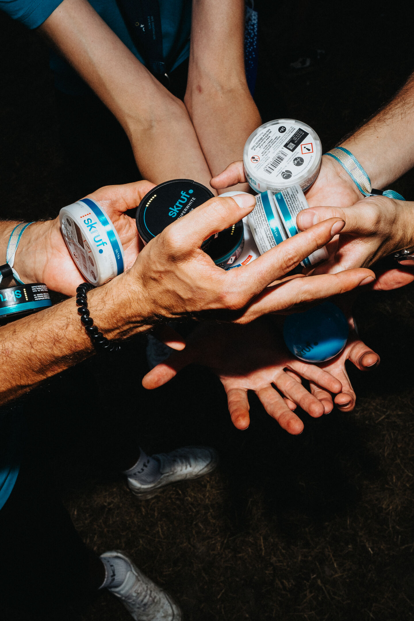 Mehrere Hände halten und stapeln runde skruf.-Dosen bei Nachtlicht auf einem Festivalgelände.