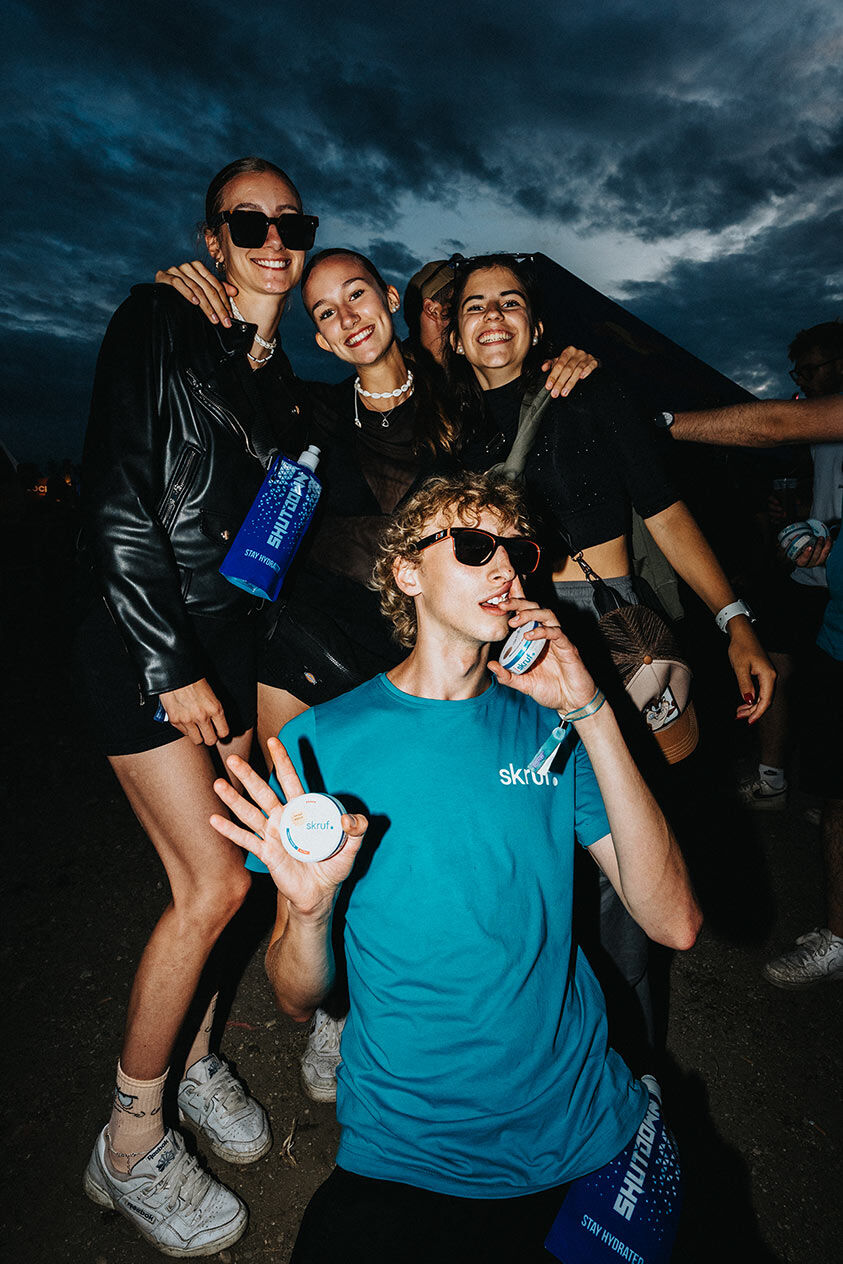 Vier lachende Freunde posieren nachts auf dem Shutdown Festival mit skruf Dosen und Festival-Branding unter einem dramatischen Abendhimmel.