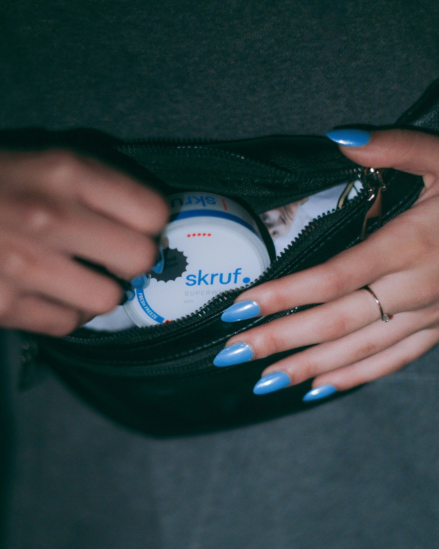 Hand mit blauen Nägeln steckt skruf.-Dose in schwarze Bauchtasche.