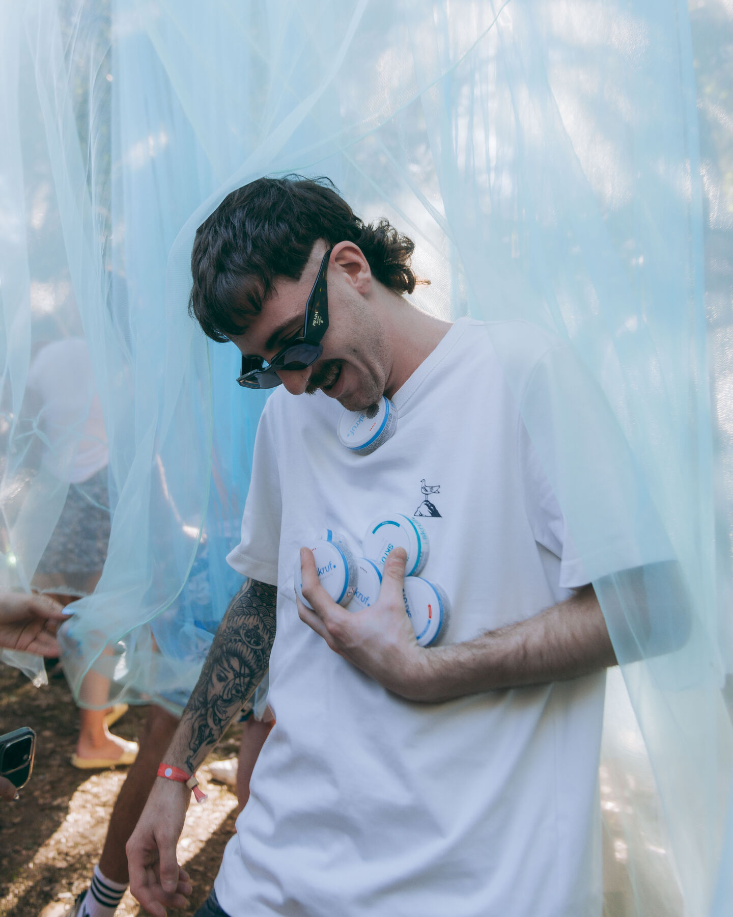 Mann mit Sonnenbrille lacht und hält mehrere skruf.-Dosen vor hellblauem Vorhang.