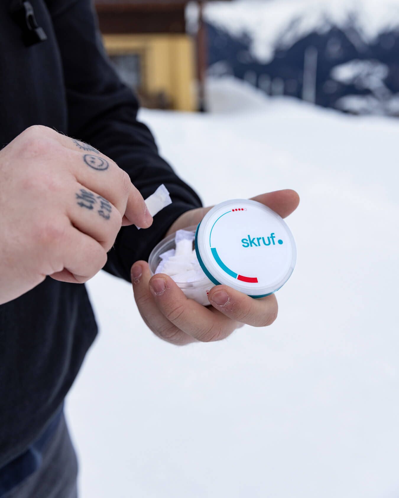 Mann mit tättowierten Fingern nimmt einen Nikotinbeutel aus einer skruf Dose, im Hintergrund ist Schnee zu sehen