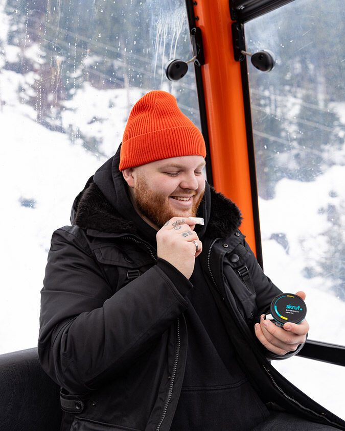 Mann in der Gondel mit orangener Haube hält eine skruf Nikotinbeutel in der einen, eine Dose in der anderen Hand