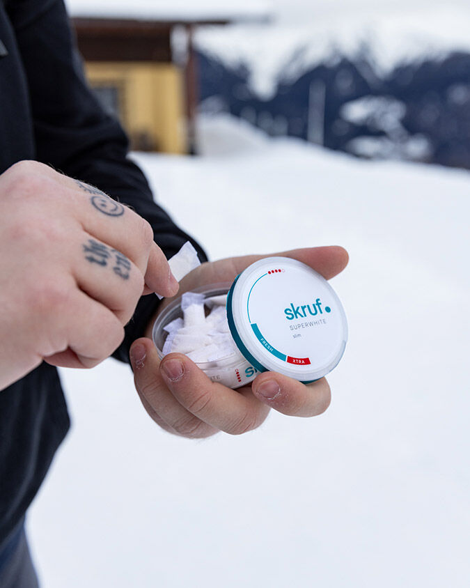 Mann mit tättowierten Fingern nimmt einen Nikotinbeutel aus einer skruf Dose, im Hintergrund ist Schnee zu sehen