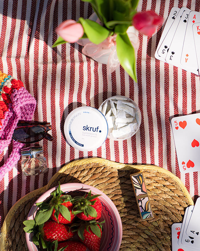 Offene Dose mit skruf Slim Pouches auf einer Picknickdecke neben Erdbeeren, Spielkarten und Sonnenbrille.
