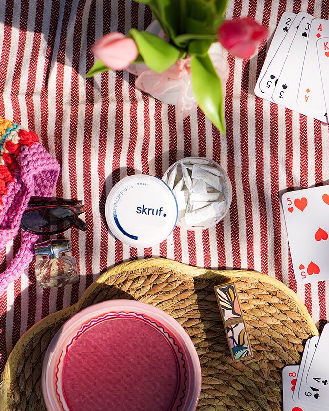 Offene Dose mit skruf Slim Pouches auf einer Picknickdecke neben Erdbeeren, Spielkarten und Sonnenbrille.