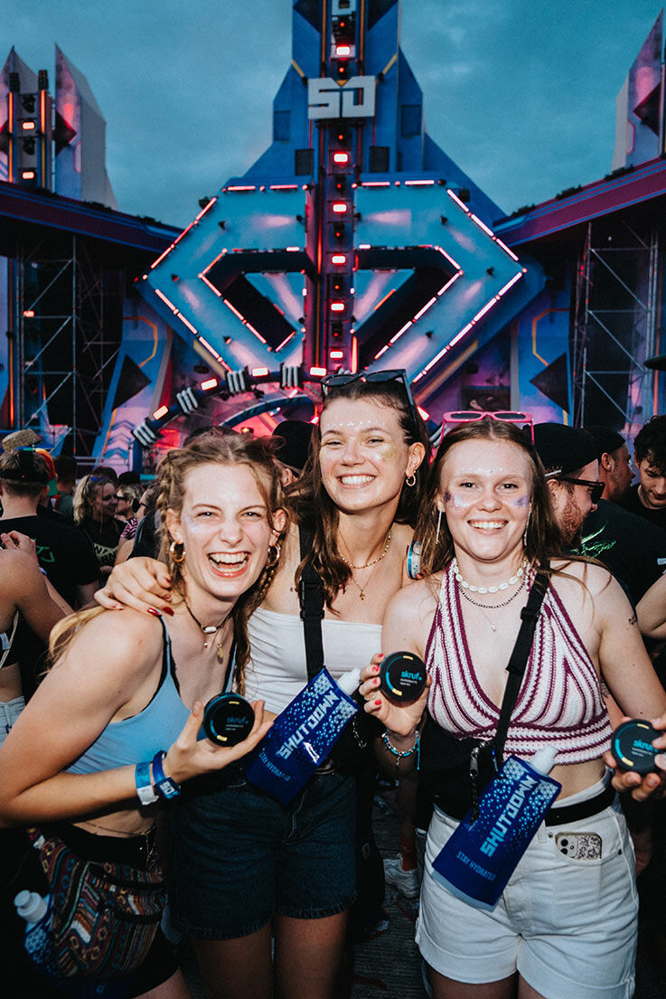 Drei lachende Frauen beim Shutdown Festival halten stolz ihre skruf Dosen in die Kamera, im Hintergrund die beleuchtete Hauptbühne.