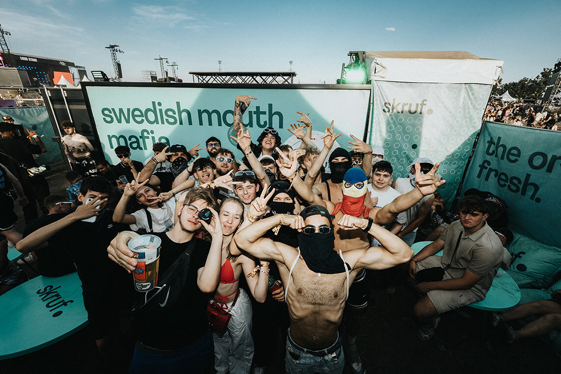 Eine große Gruppe junger Leute feiert ausgelassen am skruf Festivalstand vor einem Banner mit der Aufschrift 'Swedish Mouth Mafia'