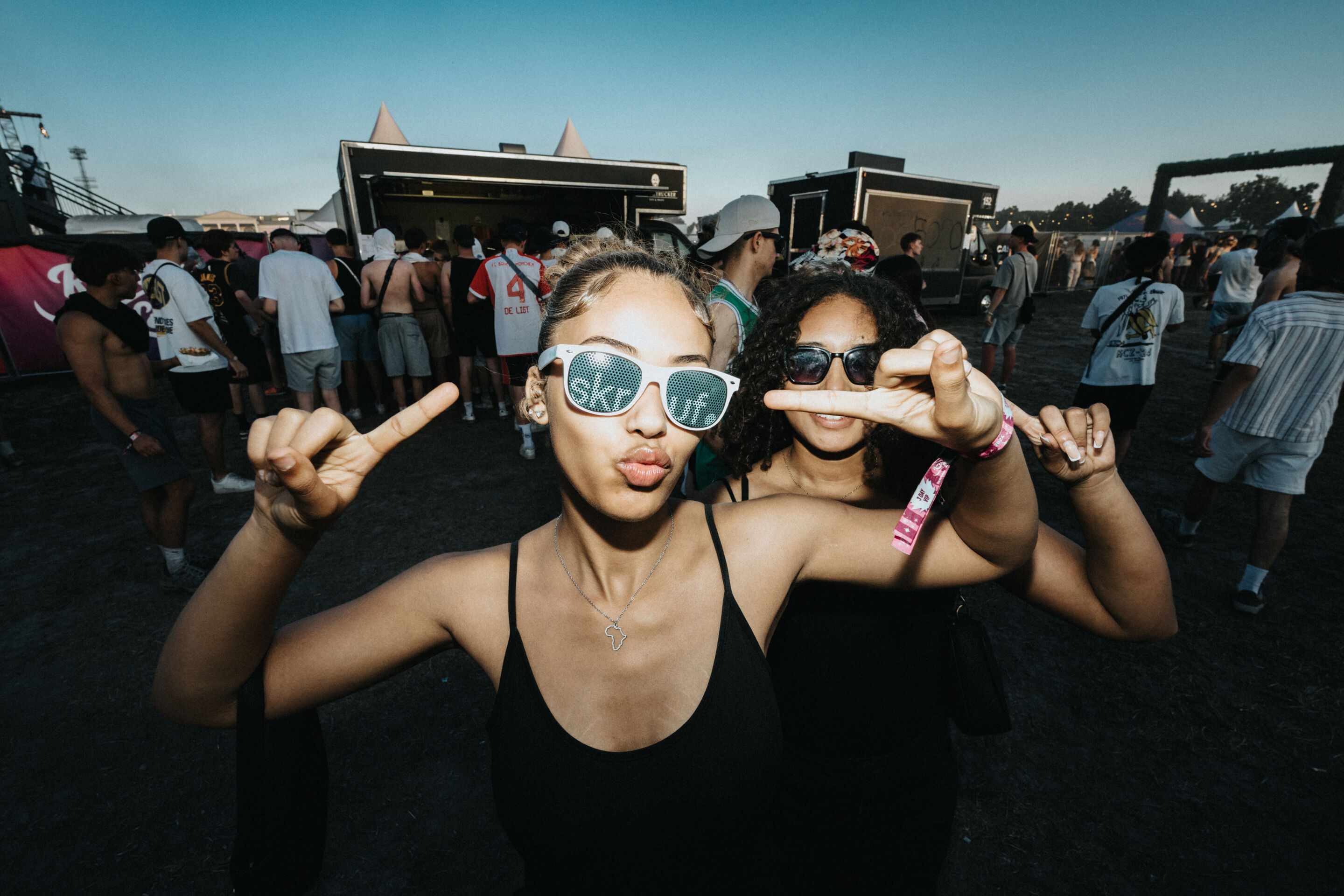 Zwei junge Frauen posieren auf einem Festivalgelände, eine trägt skruf.-Sonnenbrille und zeigt Handzeichen.