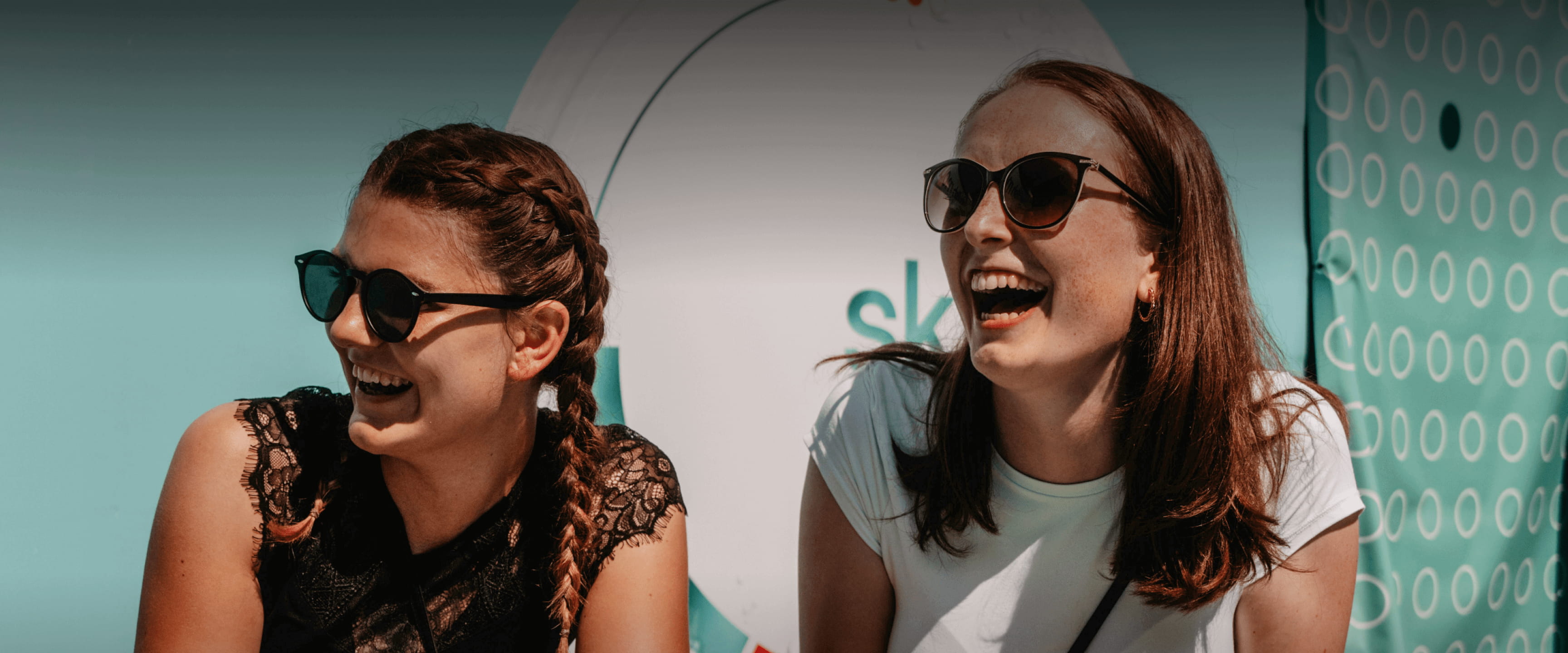 Zwei junge Frauen lachen ausgelassen und entspannt vor einem hellblauen skruf Event-Hintergrund in der Sonne.