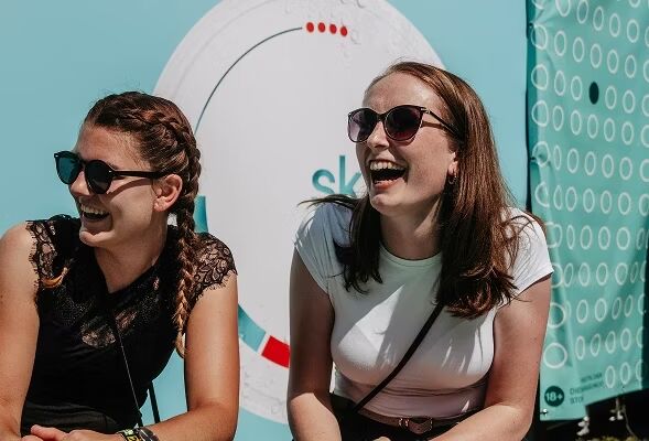 Zwei junge Frauen lachen ausgelassen und entspannt vor einem hellblauen skruf Event-Hintergrund in der Sonne
