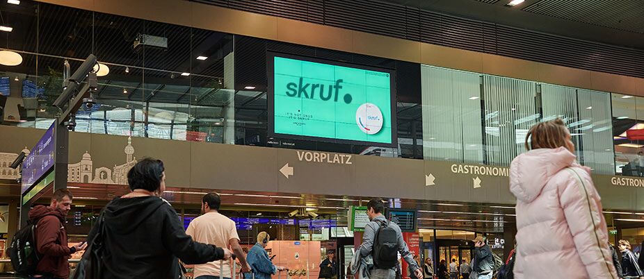 Menschen vor einer digitalen skruf Werbetafel in einer Bahnhofshalle.