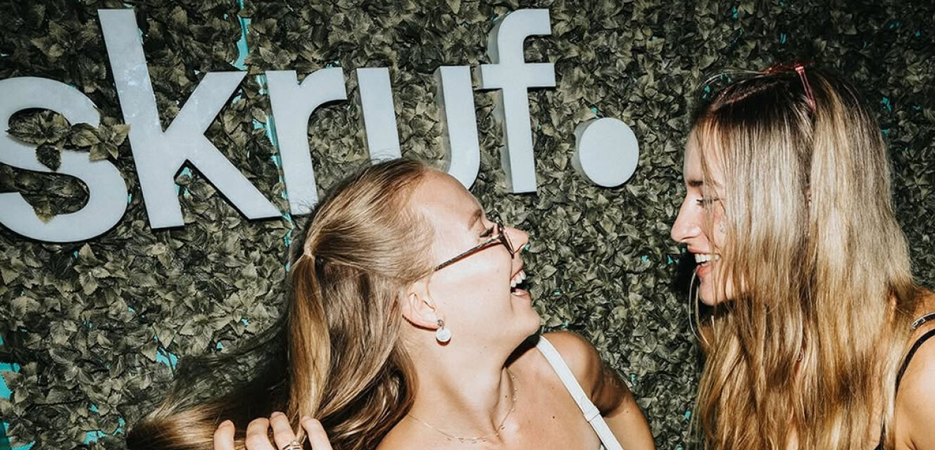 Zwei lachende Frauen vor einer Wand mit dem weißen Schriftzug „skruf.“ aus großen Buchstaben.