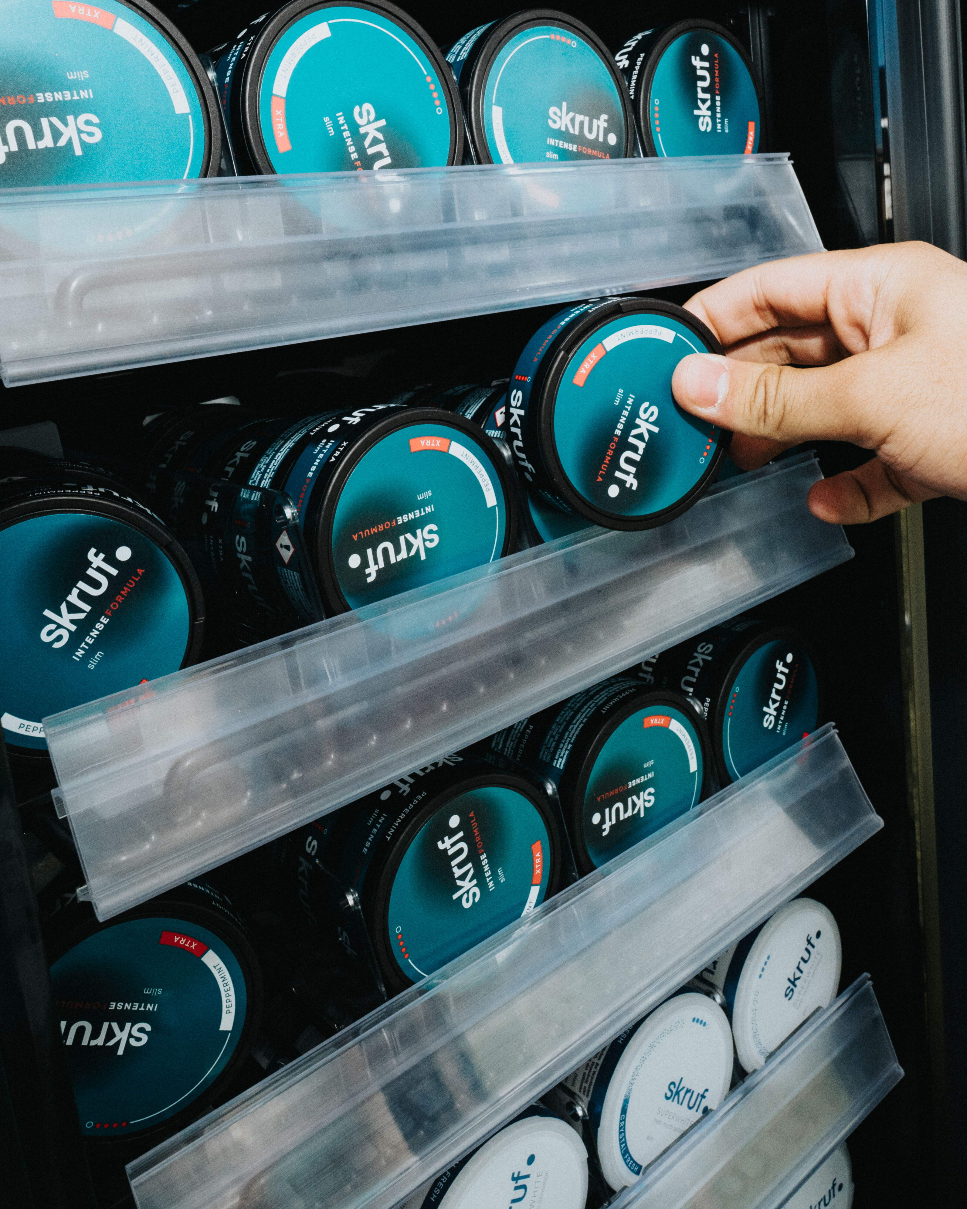 Hand greift nach einer Dose Skruf Intense Formula Slim Nikotinbeutel im Verkaufsregal mit mehreren gestapelten Skruf Dosen in verschiedenen Sorten.