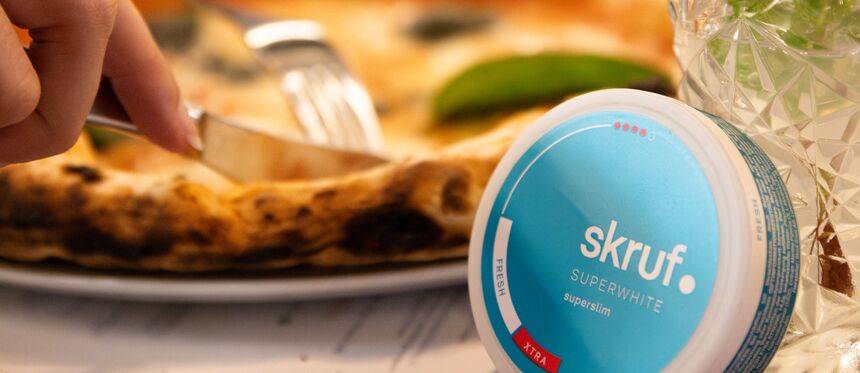 Blaue skruf.-Superwhite-Dose liegt neben Pizza und Getränk auf einem Tisch.