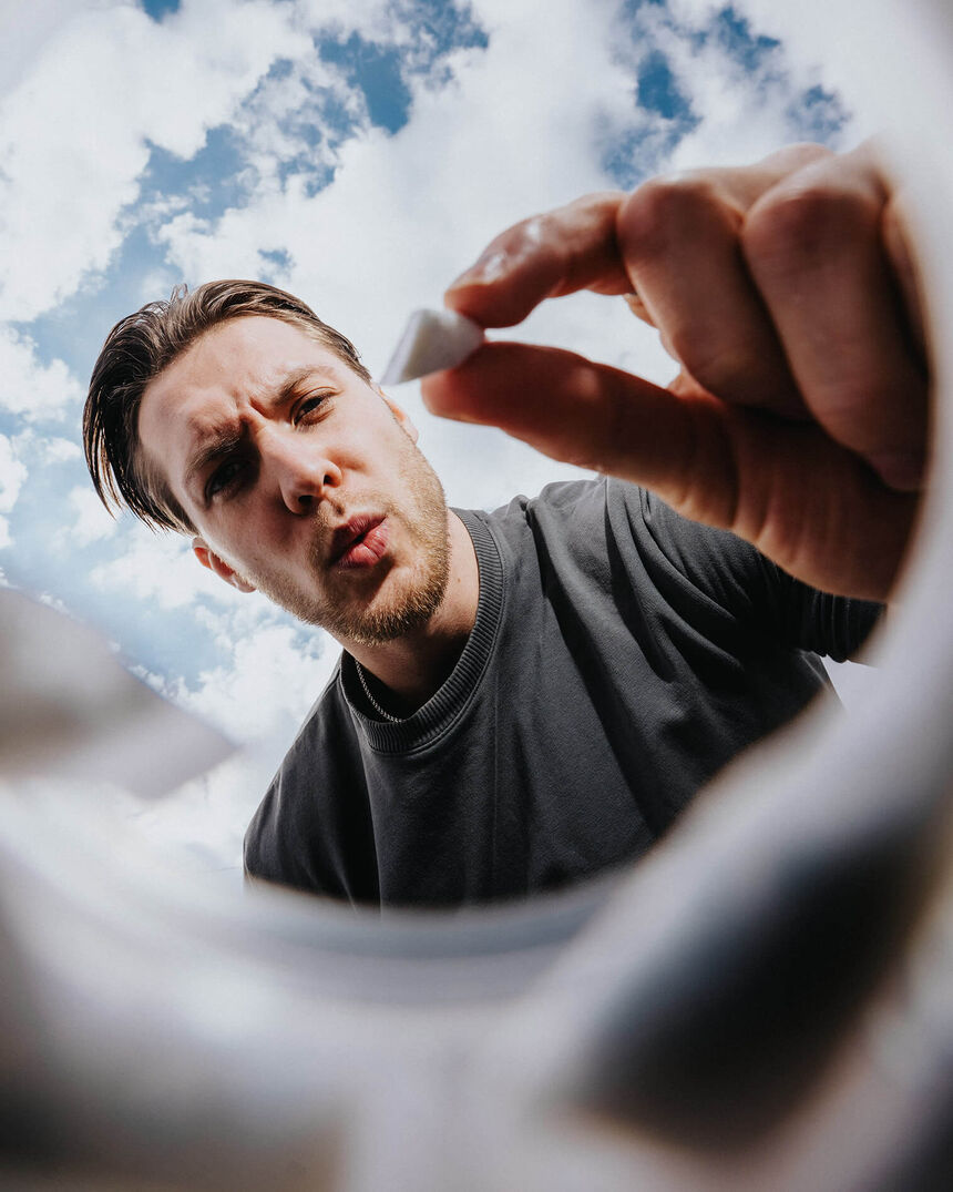 Junger Mann blickt von oben in einen Behälter und hält einen weißen Nikotinbeutel zwischen Daumen und Zeigefinger vor blauem Himmel.